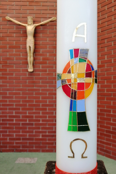 Osterkerze im Fordergrund mit Alpha, Omega und einem regenbogenfarbenen Kreuz, im Hintergrund der segnende Jesus an der Altarwand.